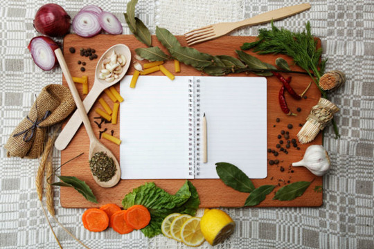 Recipe book with blank page on top of a cutting board with an array of herbs and such